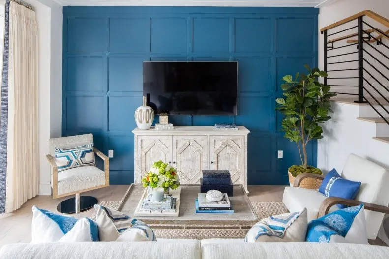 Living room with blue paneled television wall, pale low cabinet, pale sofa, blue cushions, woven armchair, rectangular table and stair rail at the side.
