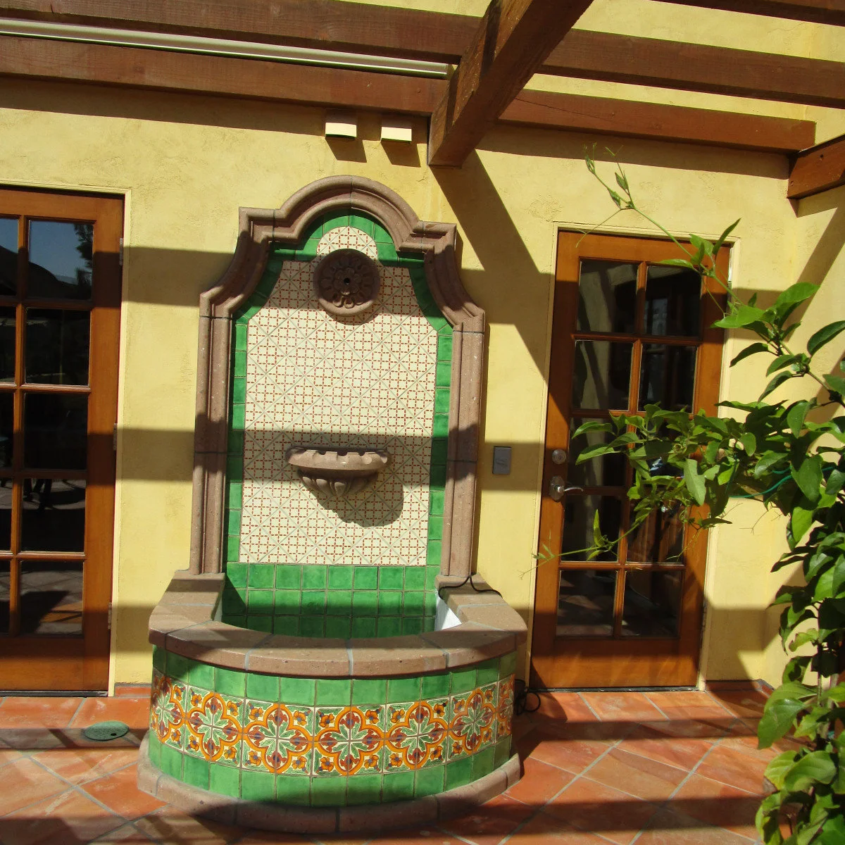 Green and patterned tile wall fountain with a shaped surround between wood-framed doors on a yellow courtyard wall.