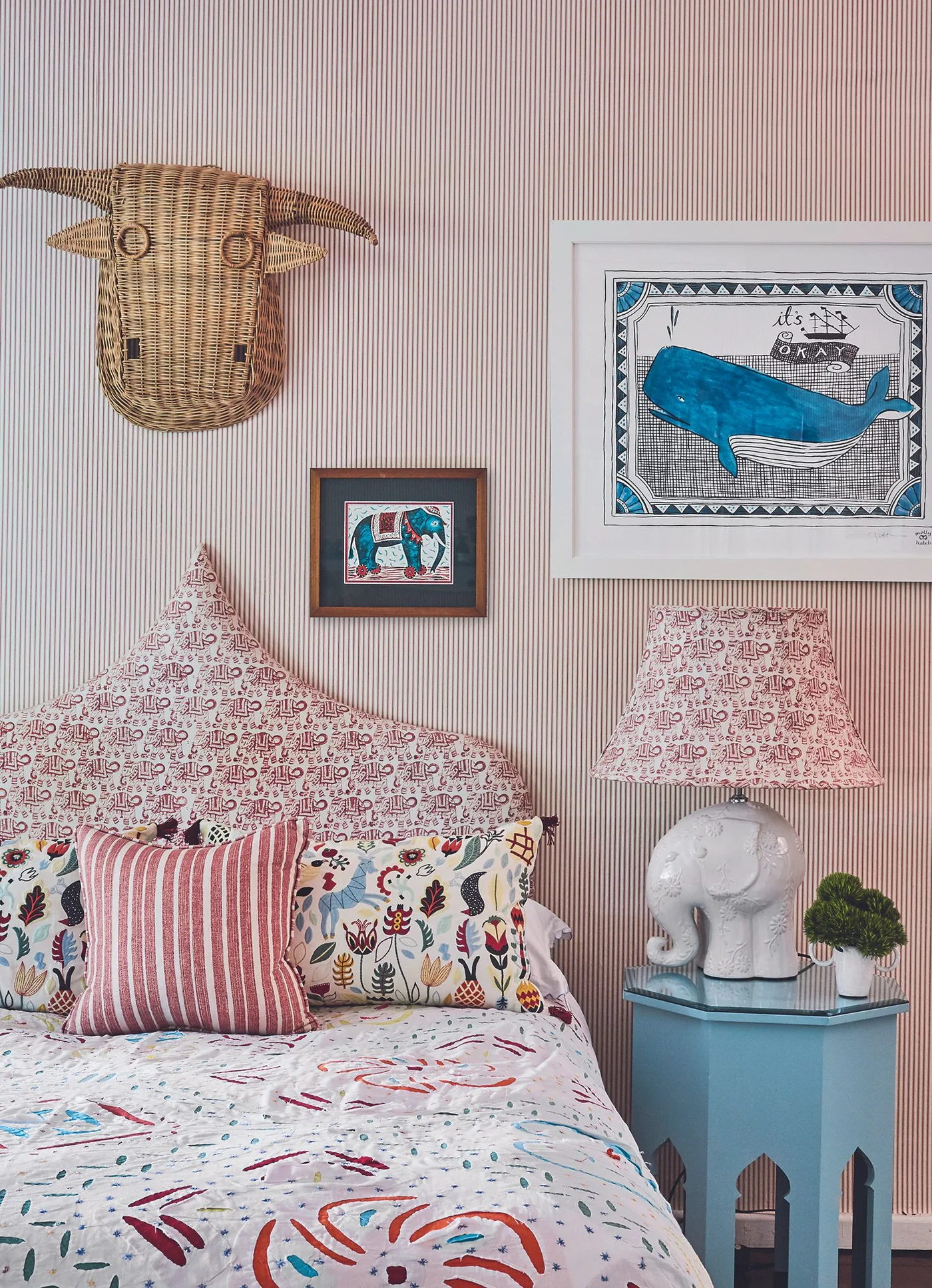 Bedroom with narrow pink striped wall, pointed patterned headboard, printed bedding, blue bedside table and elephant-shaped lamp.