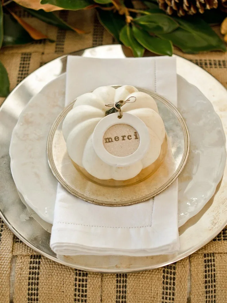 A small pale pumpkin with a round merci tag sits above a white napkin and stacked pale plates