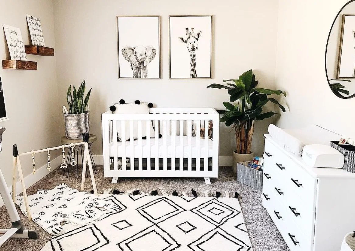 A white crib and dresser with dark handles stand beside pale walls, black-framed animal pictures, a large black-and-white geometric rug and plants