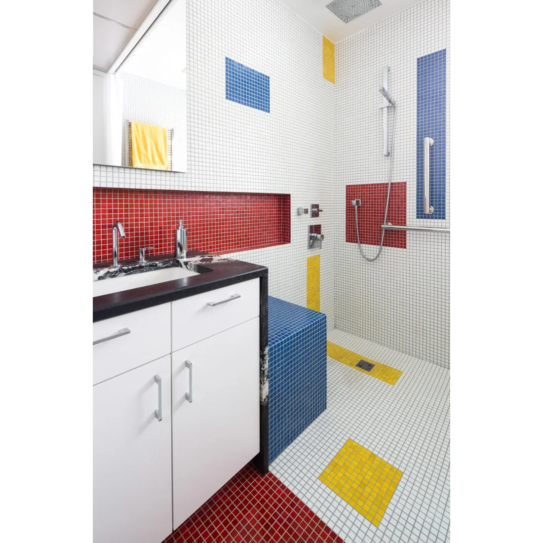 White square mosaics contain red, blue and yellow blocks across a bathroom wall, seat and floor.