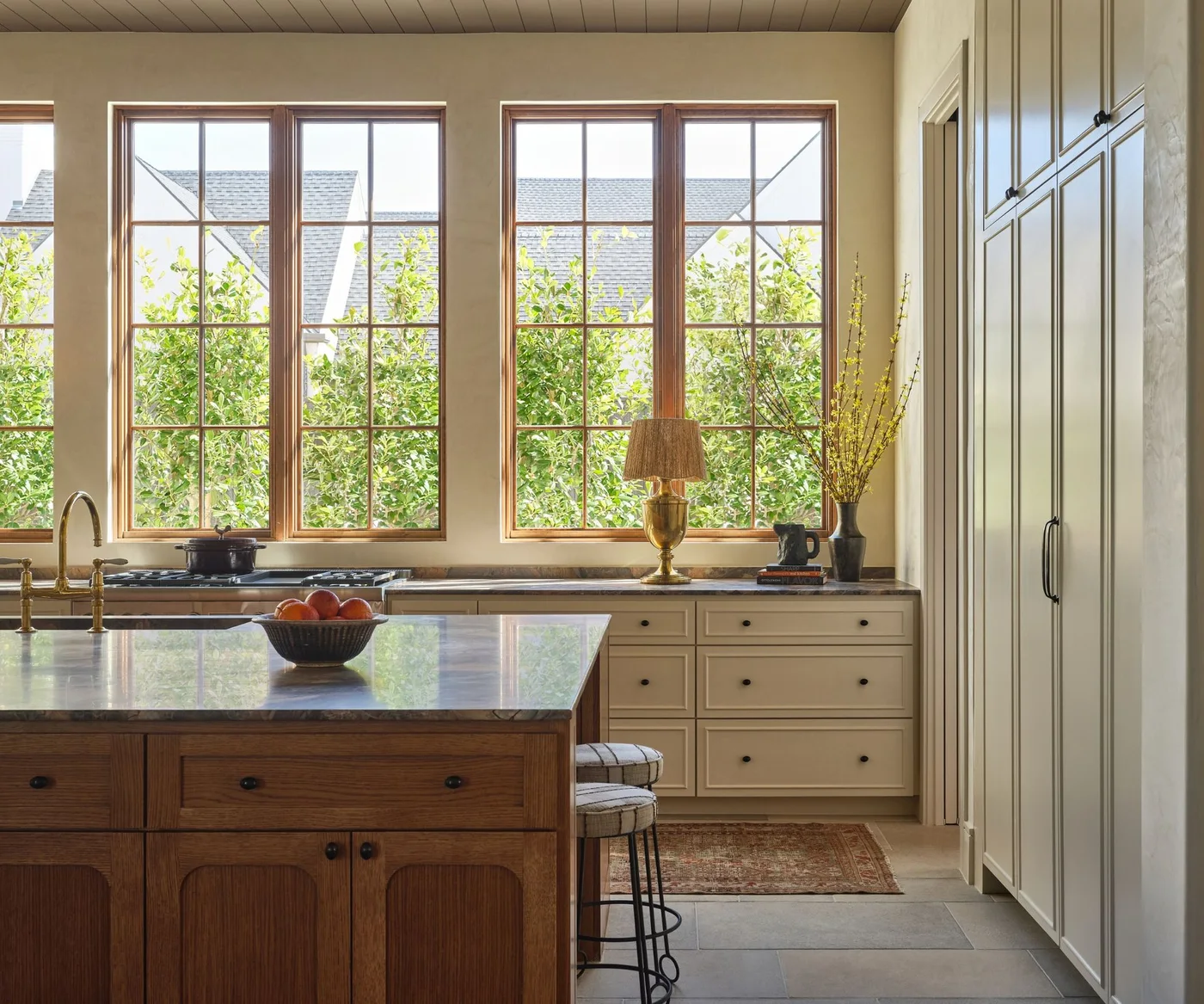 A pale off-white kitchen has broad wood-framed windows, a wood island, dark counters, small black cabinet knobs and a wood-paneled ceiling.