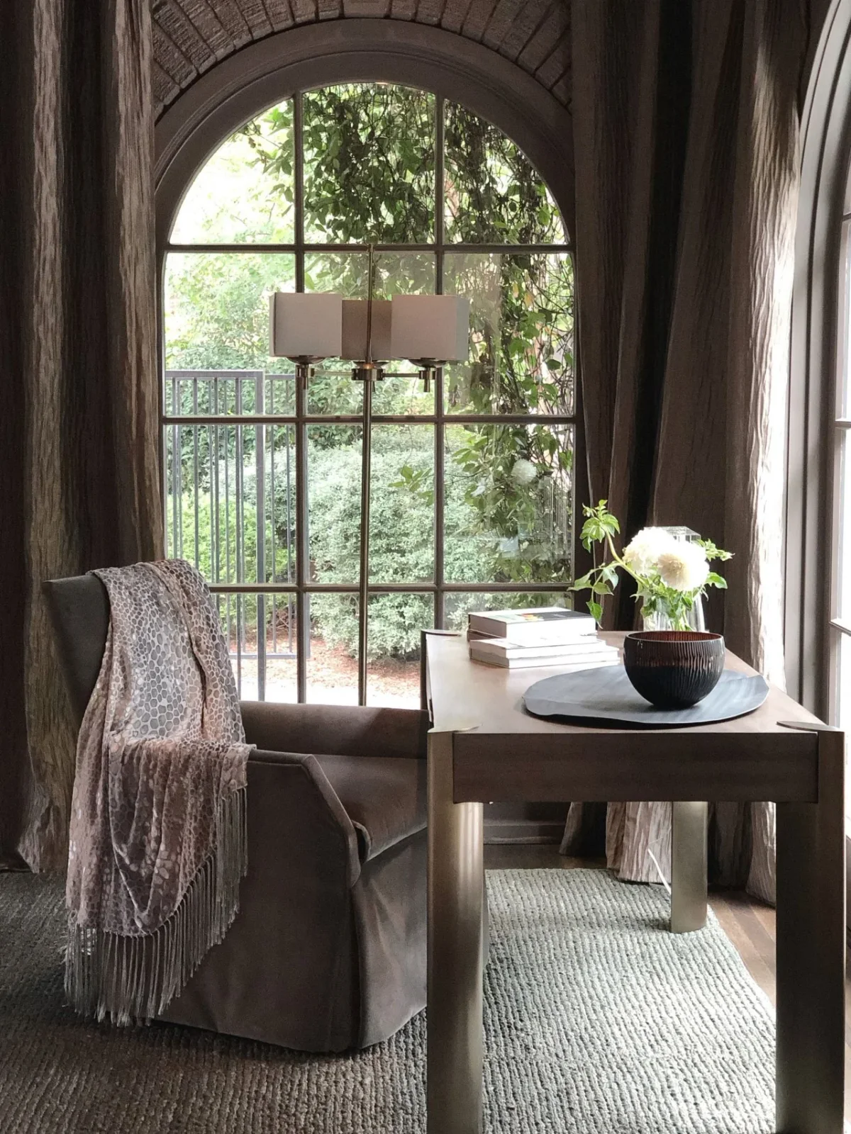 A small wood table stands beside an upholstered chair beneath an arched window, with long curtains, a hanging light and a textured rug.