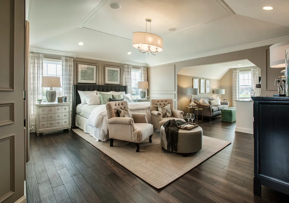 A dark bed with pale bedding and beige chairs sits on a light rug above dark wood flooring