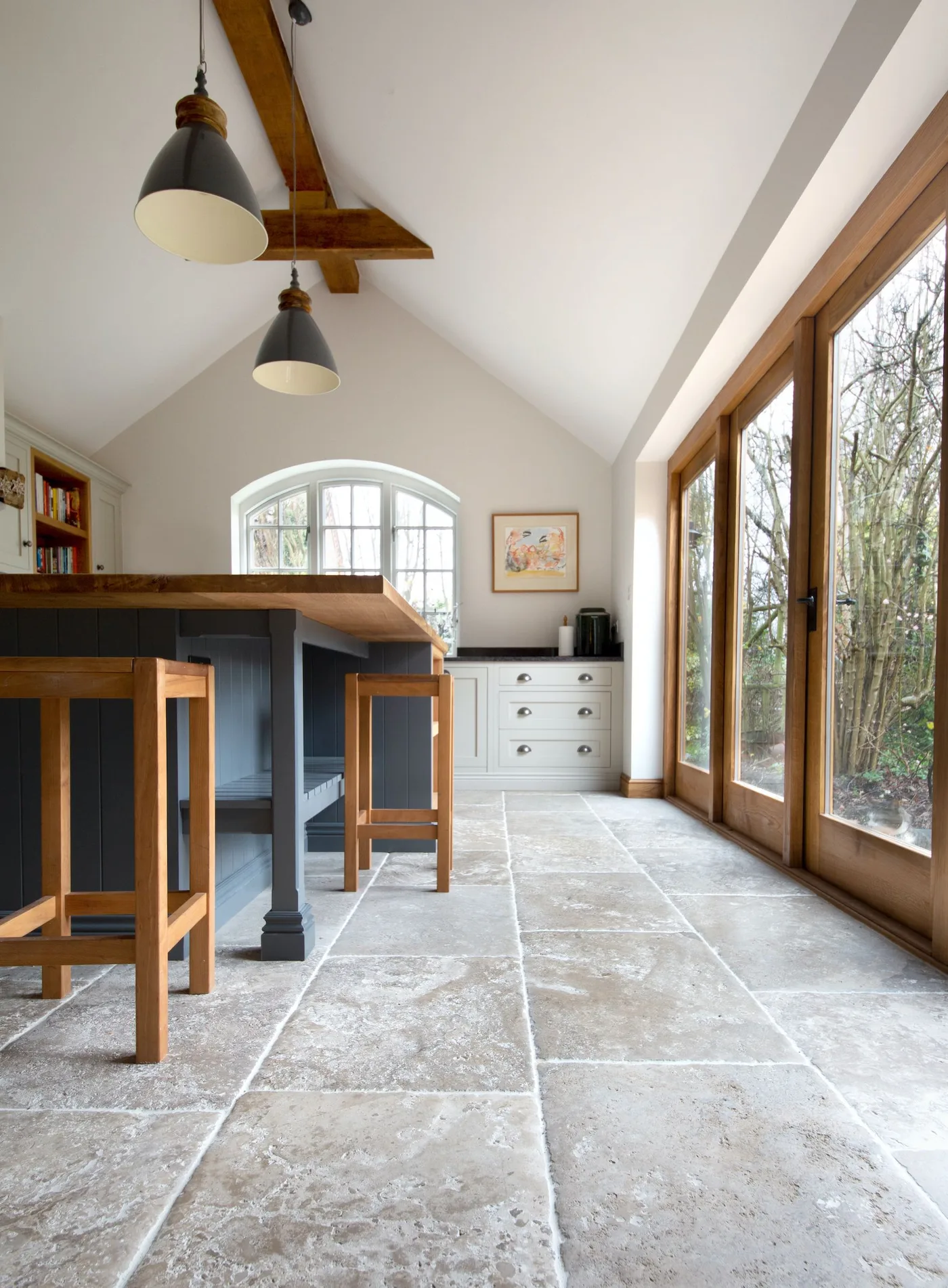 Country kitchen with dark island, wooden stools, pale rectangular floor, high white ceiling with exposed beam and glazed wooden garden doors.