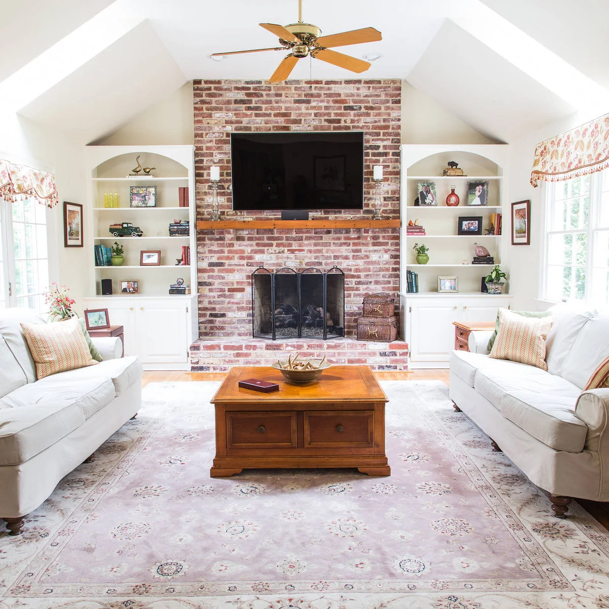 A full-height mixed red brick fireplace stands between pale built-in shelves beneath a sloping ceiling, with a wood mantel and pale sofas.