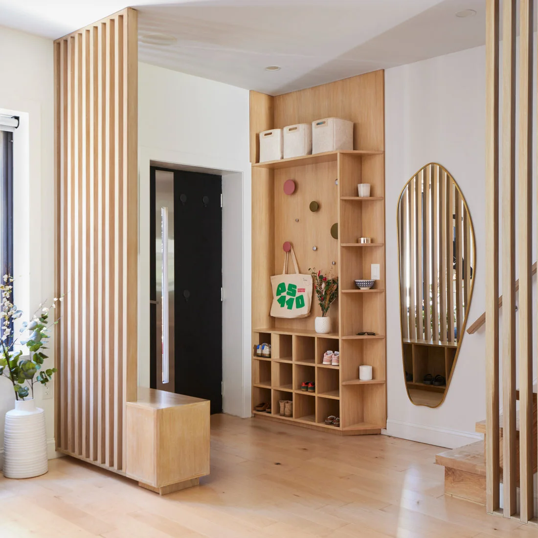 A pale wood entry unit combines shoe cubbies, colorful round hooks and small shelves beside a wood slat divider and shaped mirror.