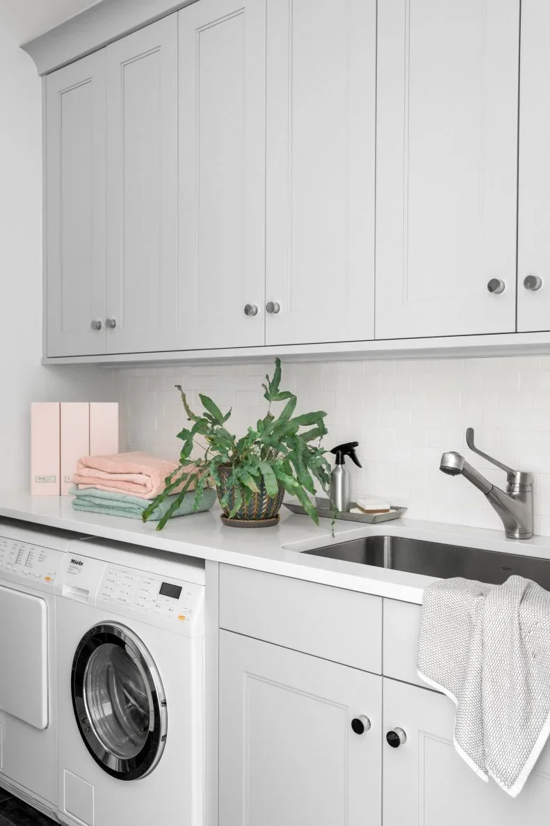 A gray laundry counter combines a metal sink, side-by-side laundry appliances, pale backsplash tiles and upper cupboards.