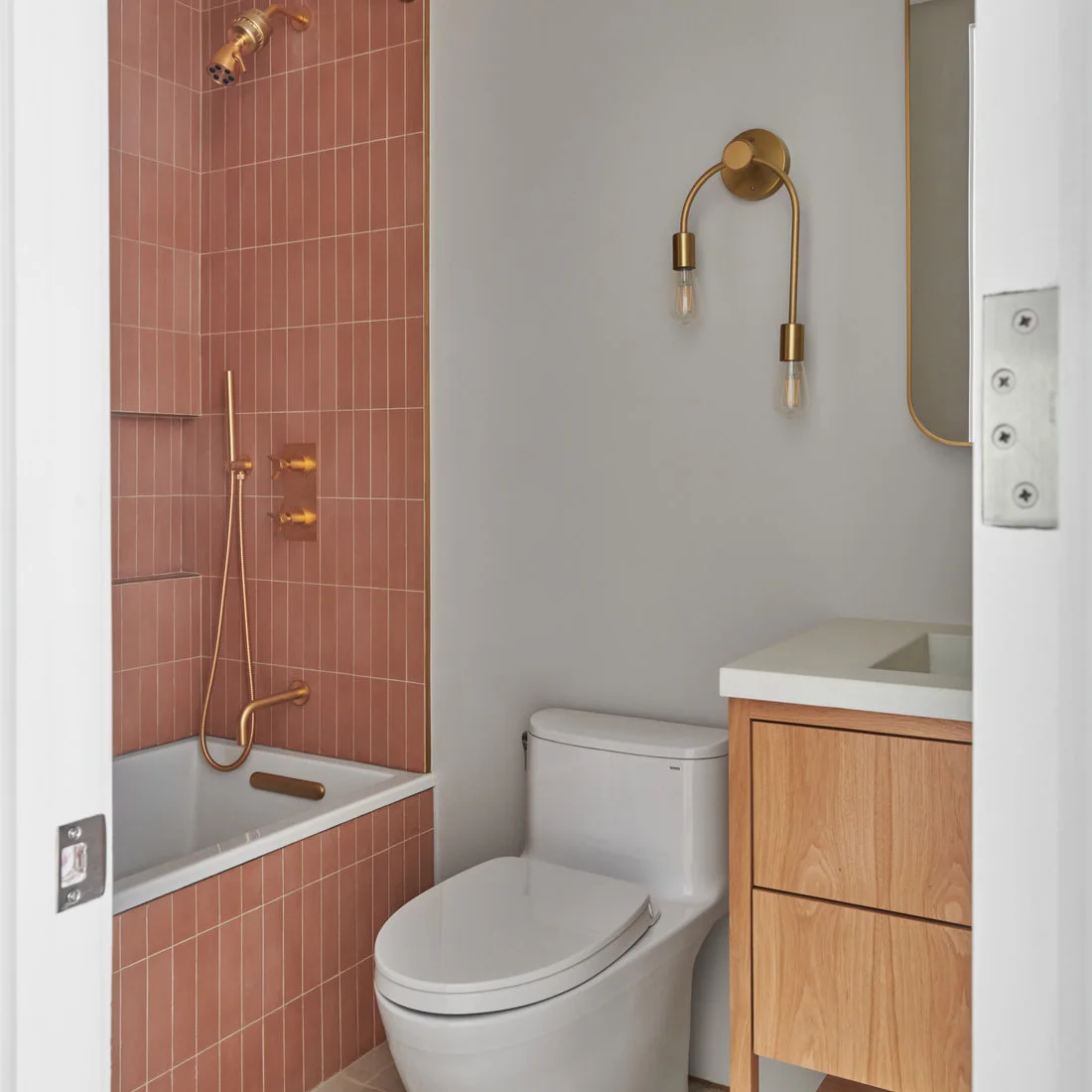 Deep pink tiles stand vertically around a white bath beside pale walls, a toilet and a wooden vanity