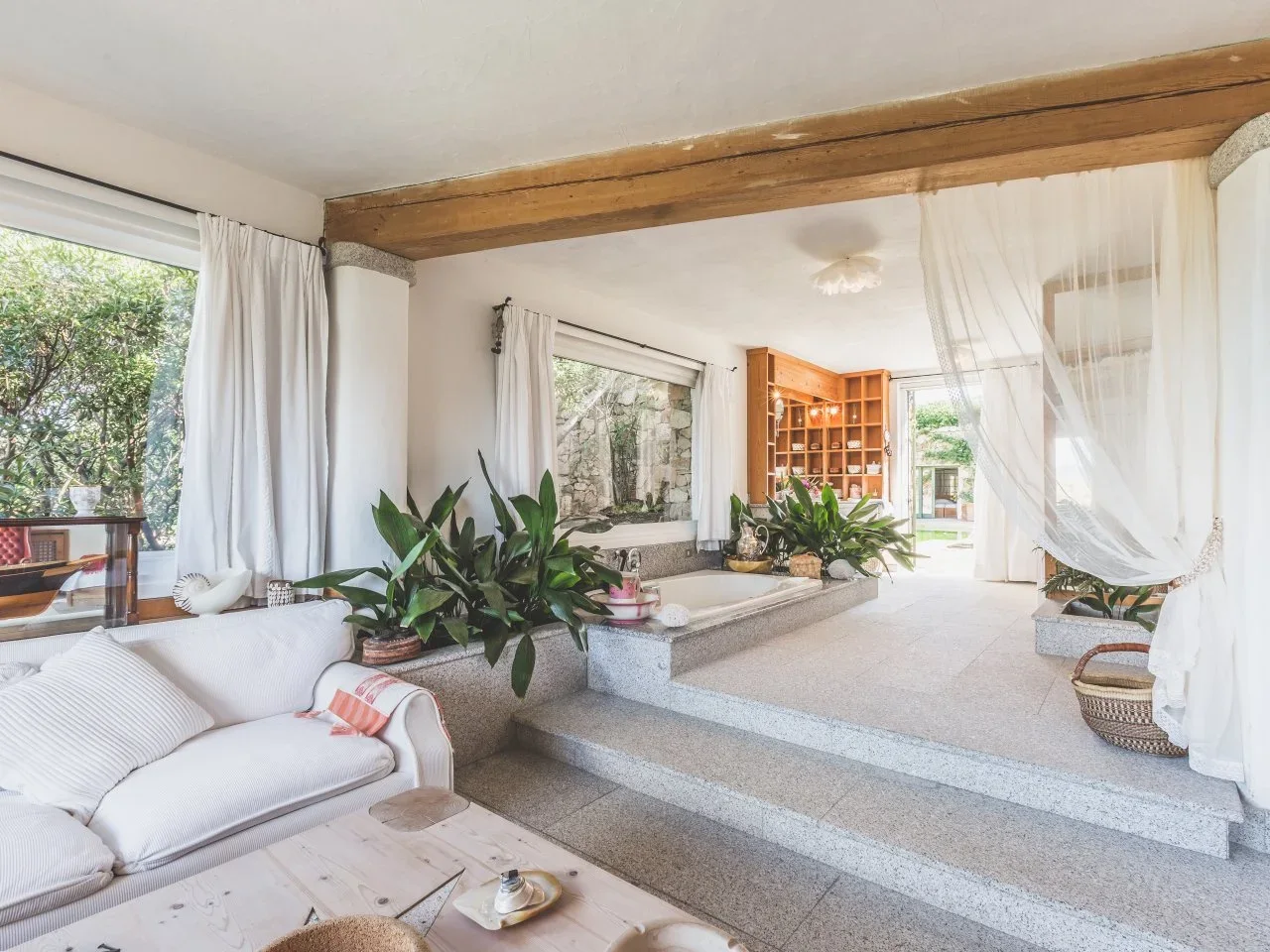 Plants border a tub within a raised pale platform beside sheer curtains, a timber beam and large windows.