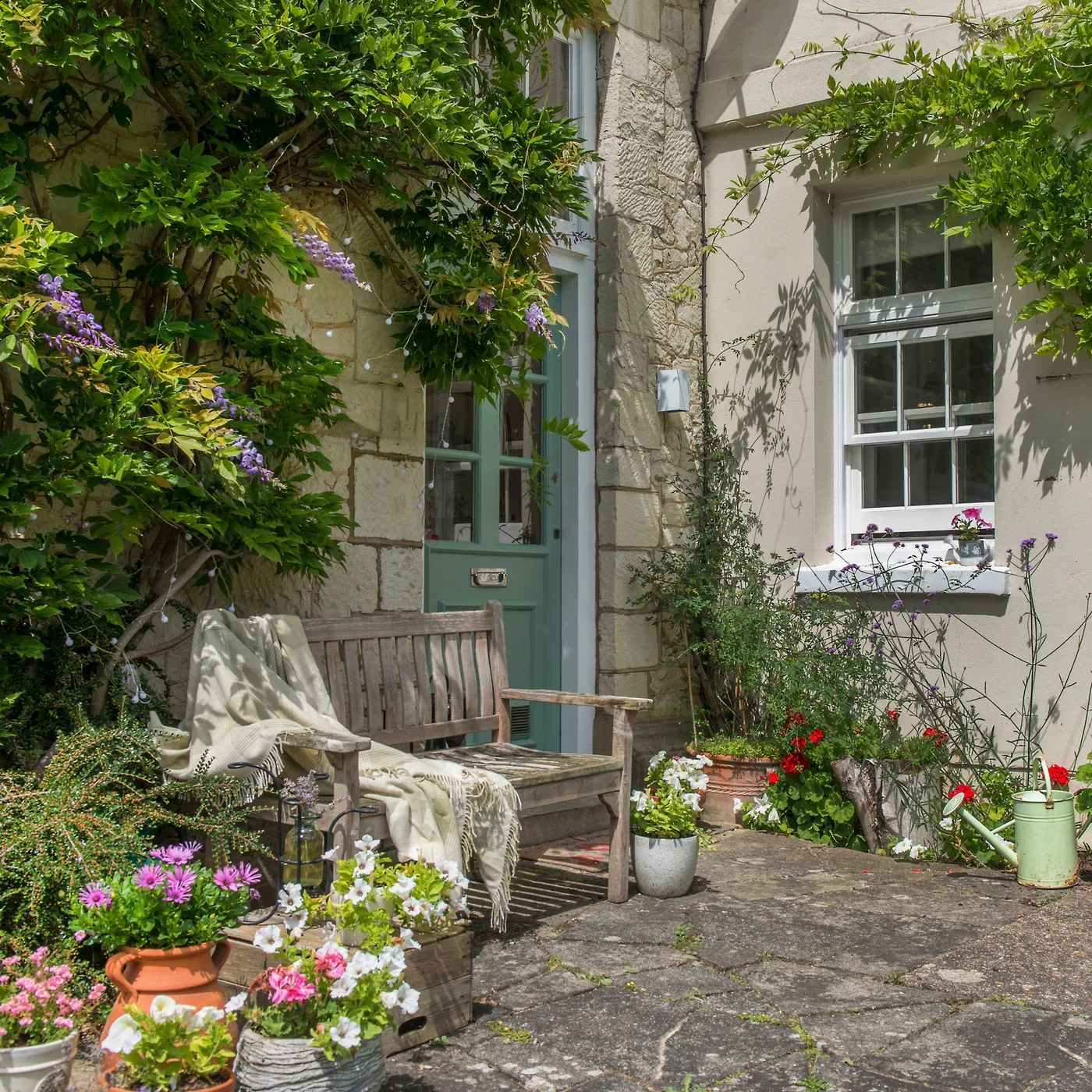 A weathered timber bench sits beside a pale house wall among flowering containers