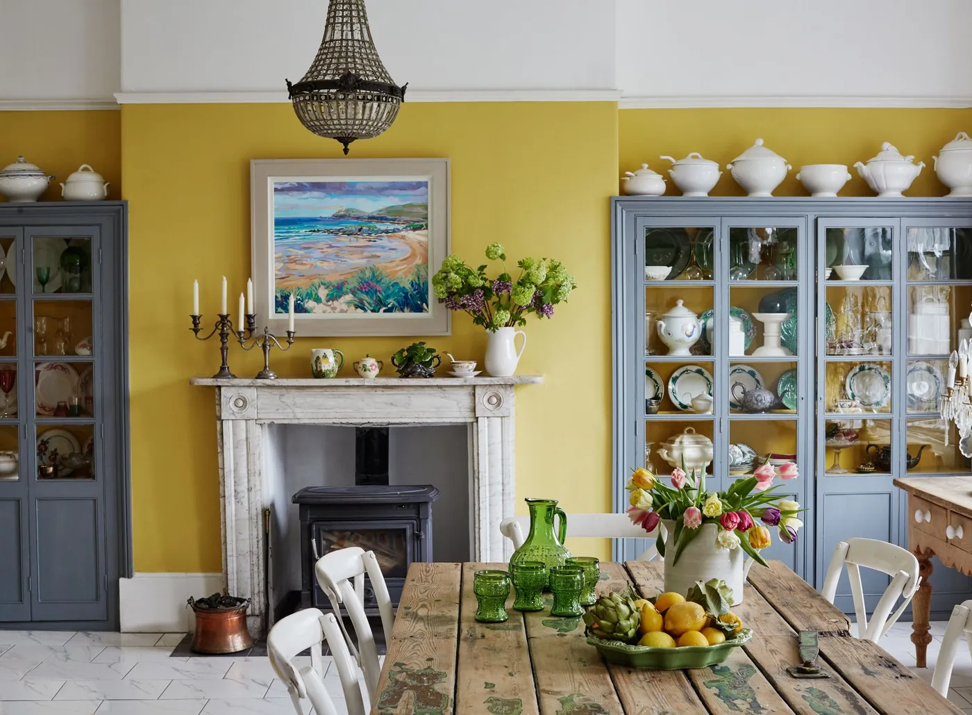 White fireplace surround with dark stove, yellow dining room walls, gray cupboards and wooden table.