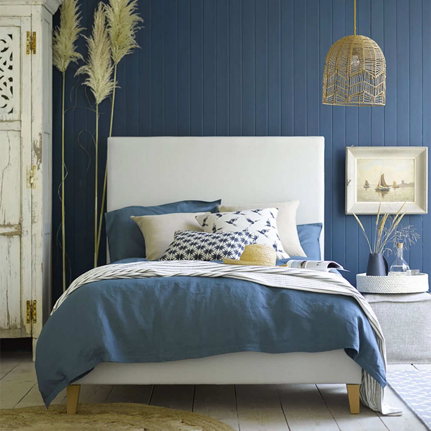 Deep blue paneled bedroom wall with a pale headboard, blue bedding, woven pendant, and distressed pale cabinet.