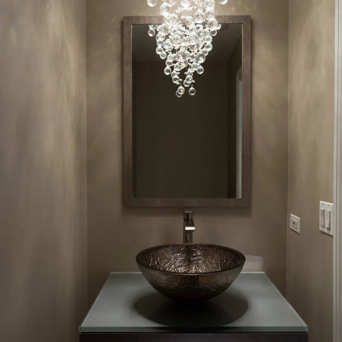A dark textured vessel basin beneath a tall rectangular mirror and a cascading glass-bubble light.