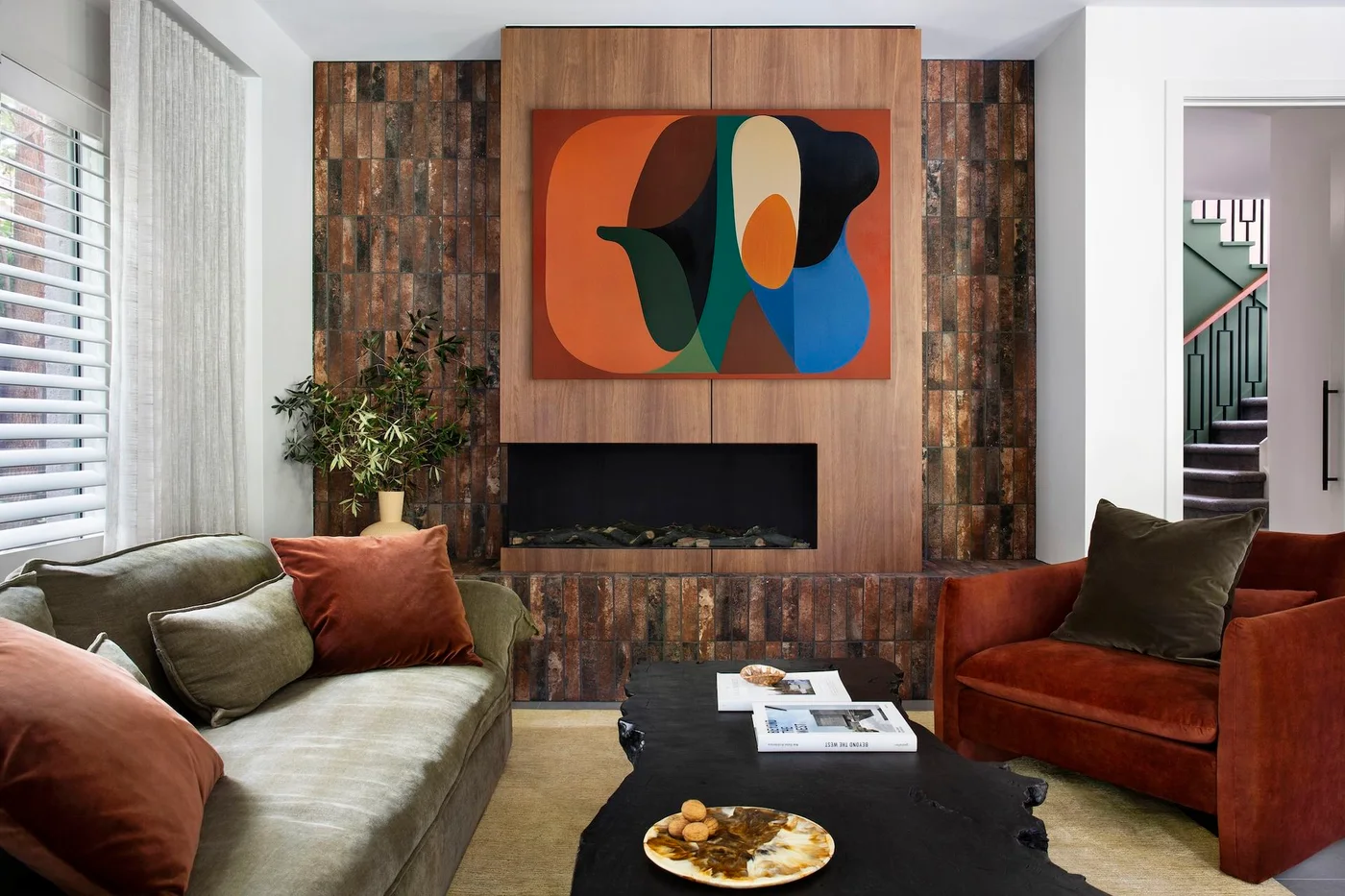 Slim brown vertical tiles flank a broad wood-toned fireplace panel with colorful artwork above, facing green and rust-colored sofas.