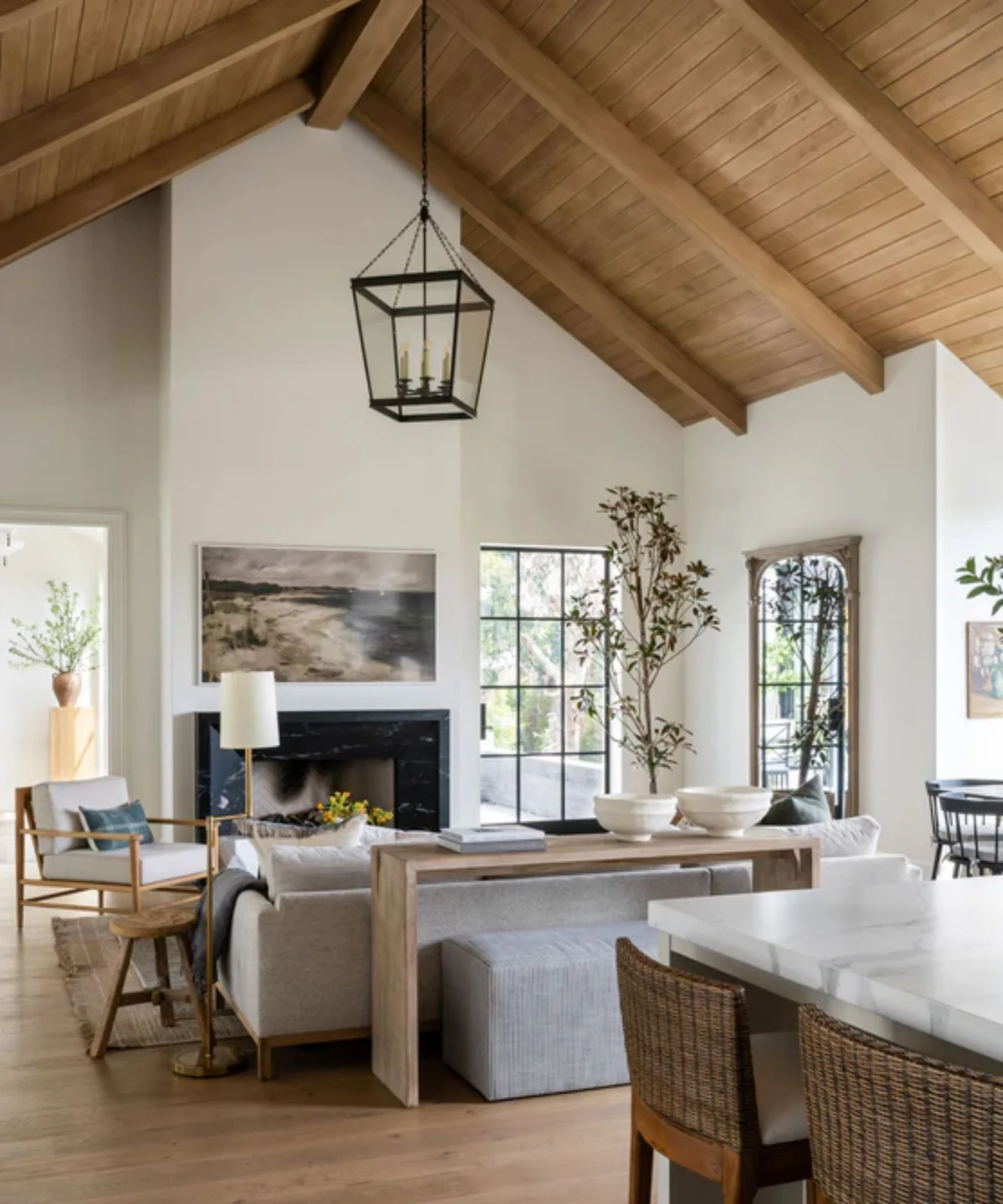 A timber console stands behind a pale sofa beneath a wood ceiling beside an open kitchen and black-framed glazing.