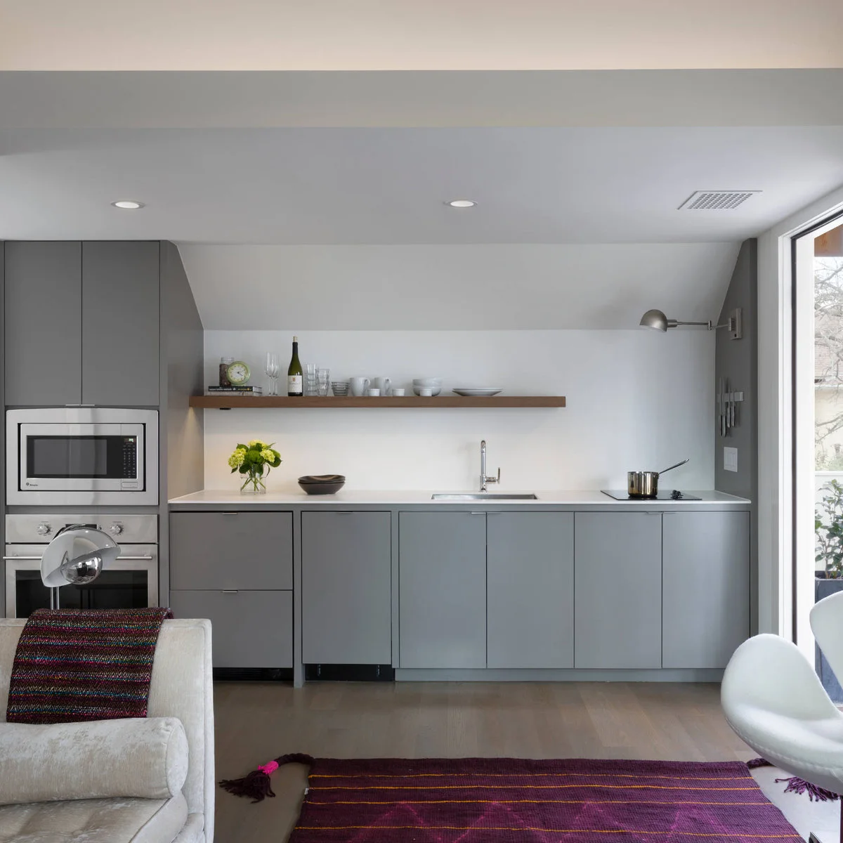 Gray one-wall kitchenette with a sink, compact cooktop, stacked appliances and one wood shelf