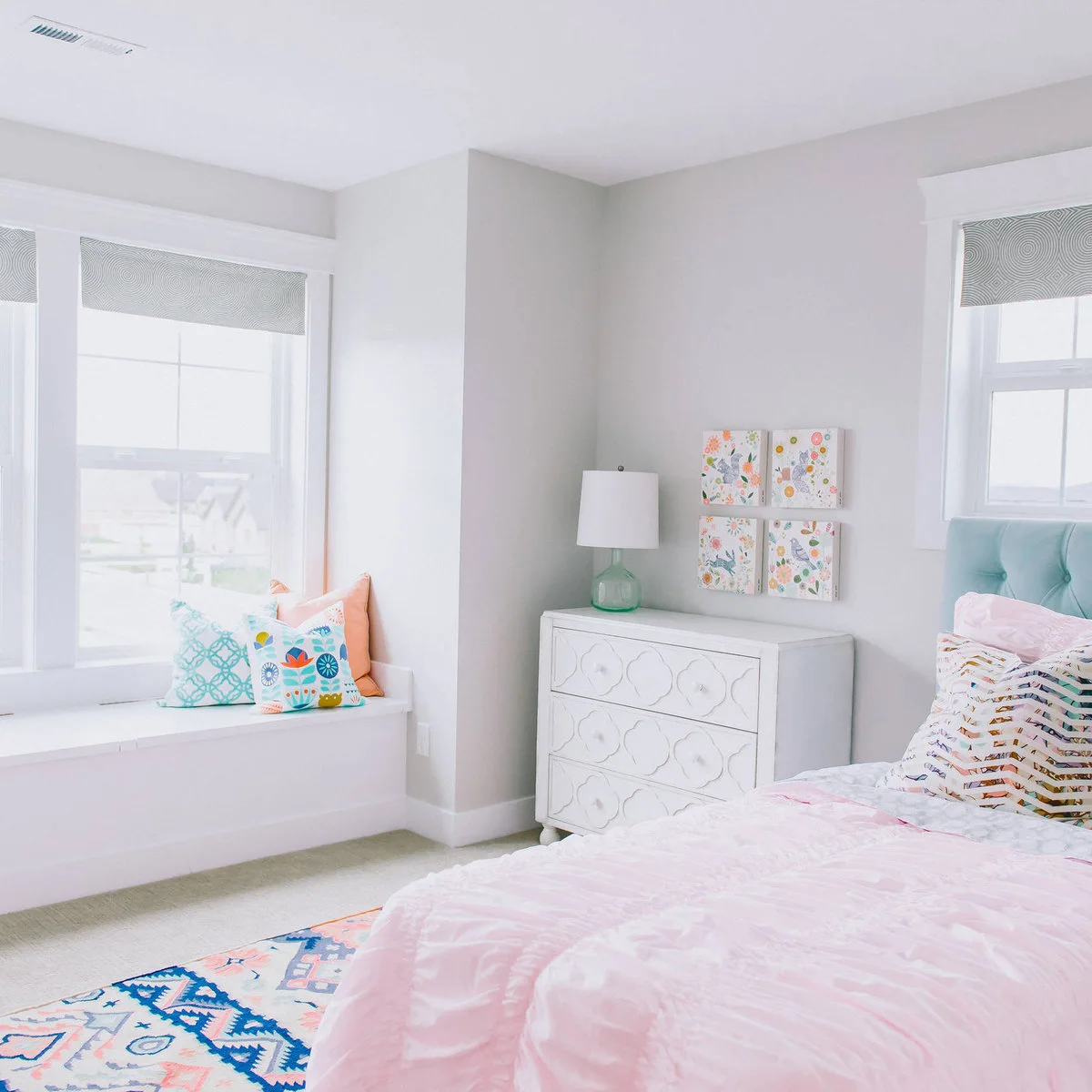 Bedroom with a pale aqua tufted headboard, pink bedding, a cushioned window bench and four floral pictures