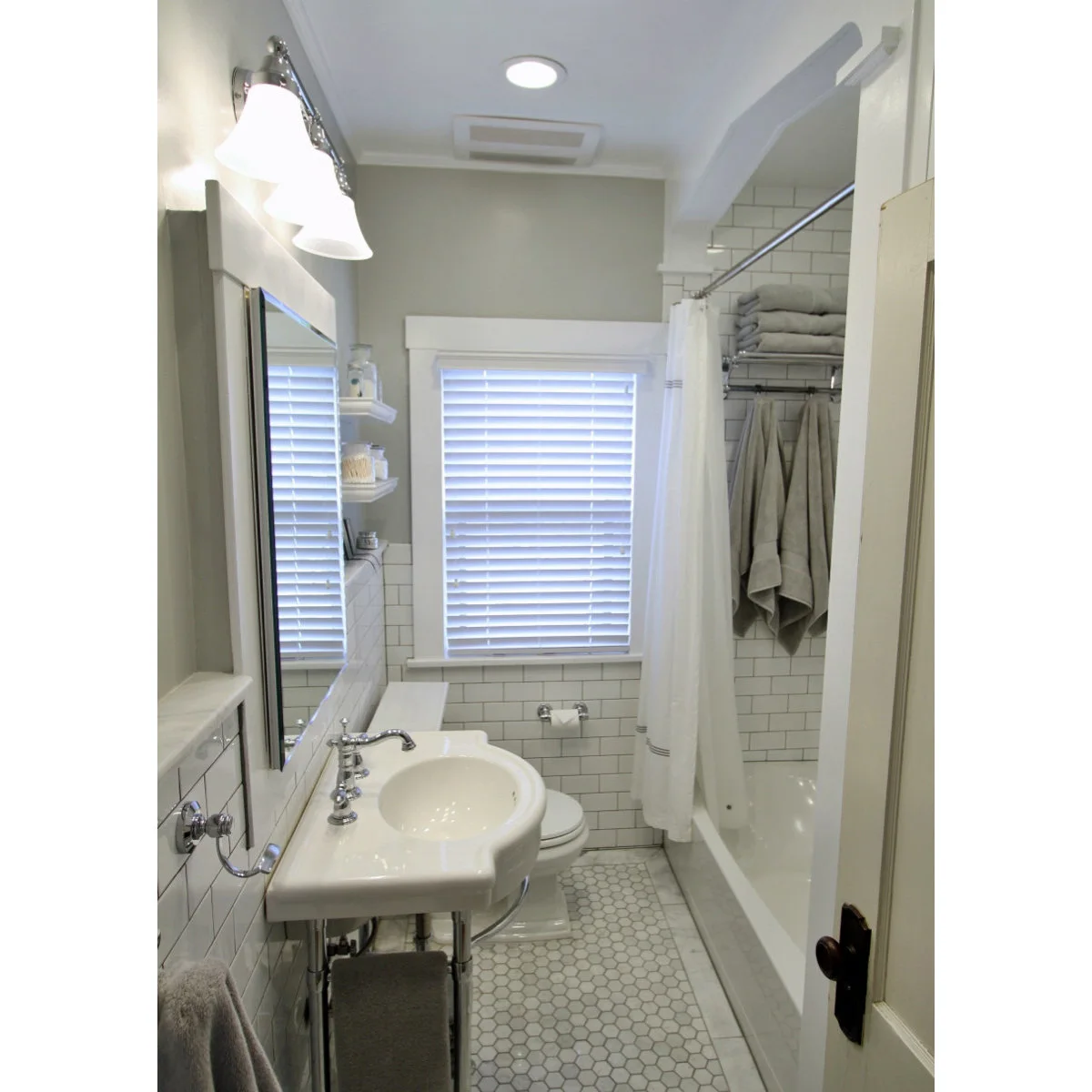 A console basin stands along a narrow bathroom with white rectangular wall tile, small hexagonal floor tiles and a white bath curtain.