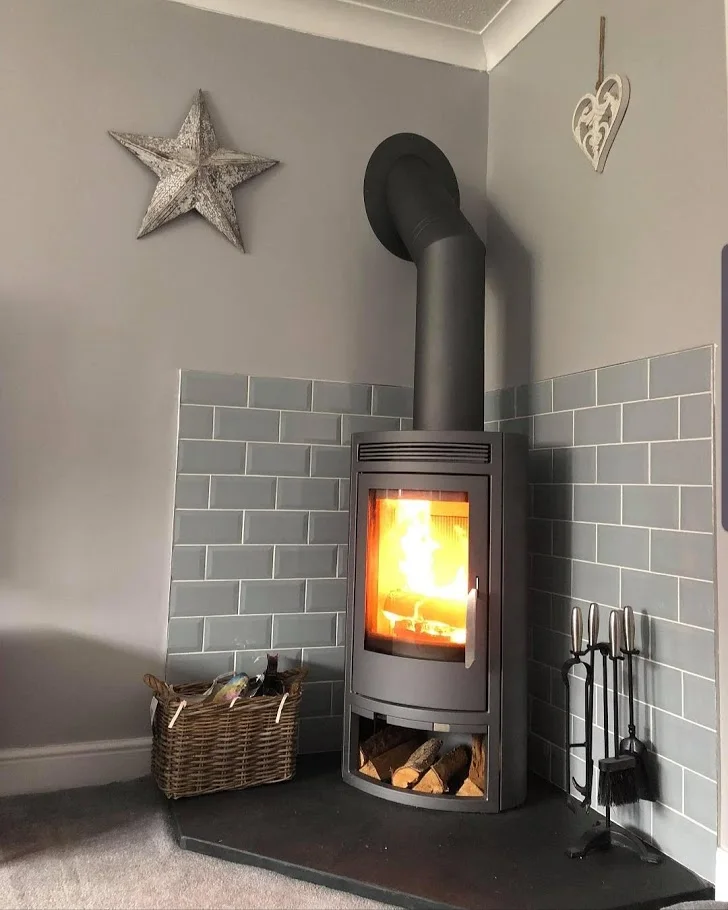 A tall gray stove stands on a dark five-sided slate hearth between tiled corner walls and a carpeted floor.
