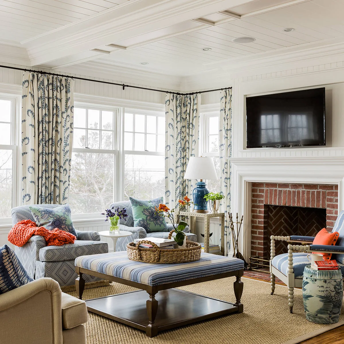 A small red brick fireplace inset sits within a pale surround beside blue patterned chairs, curtains and an upholstered ottoman.