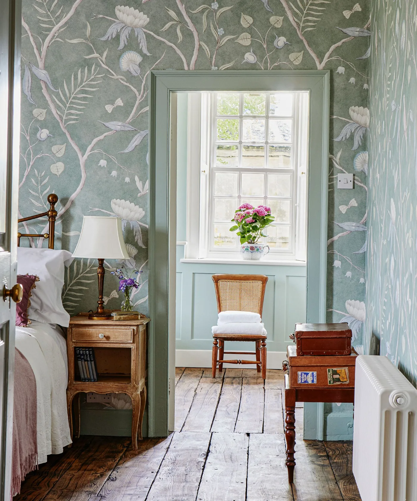 Pale green bedroom wallpaper with large white flowers around a green-trimmed window and wooden furniture.