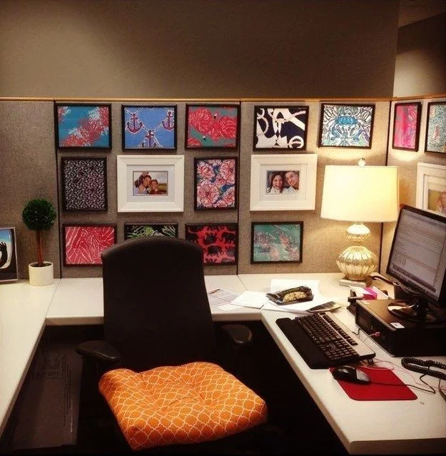 Colorful framed patterns and family photographs on cubicle panels with a clear pale desk, glass-based lamp and orange chair cushion.