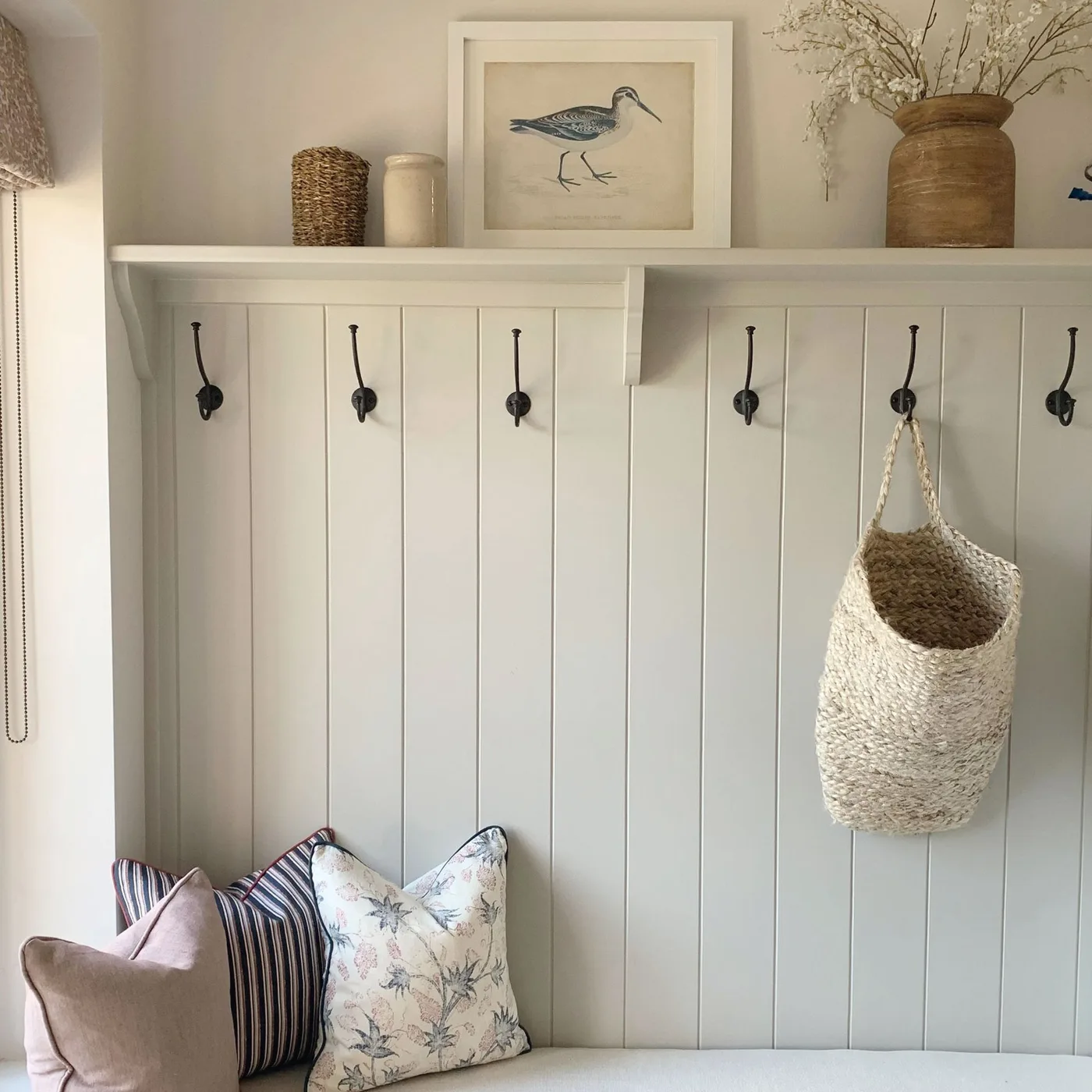 Entrance bench detail with pale vertical paneling, black coat hooks, a high shelf, mixed patterned cushions and a hanging woven basket.