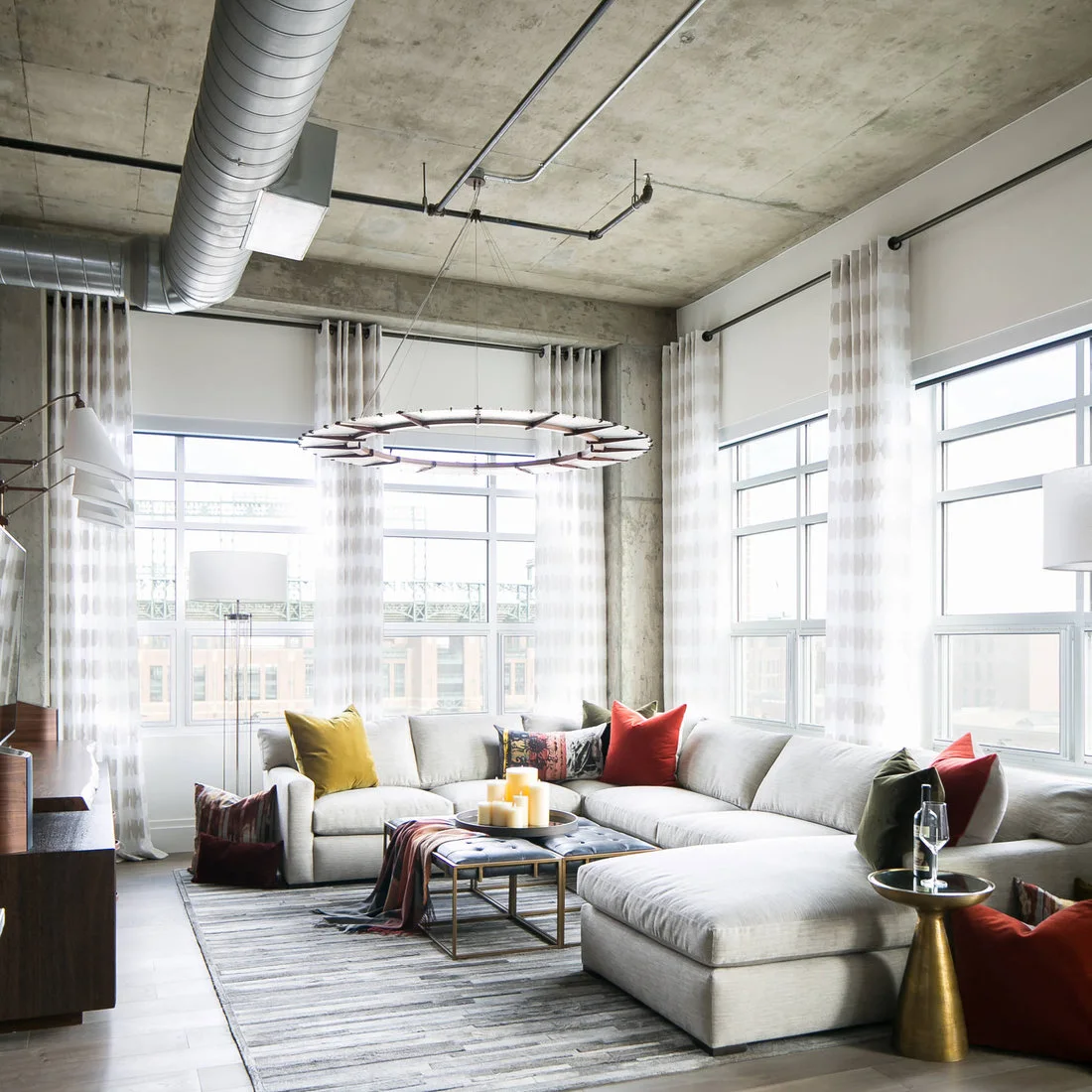 A pale wraparound sectional with colored cushions beneath exposed ducts and tall windows