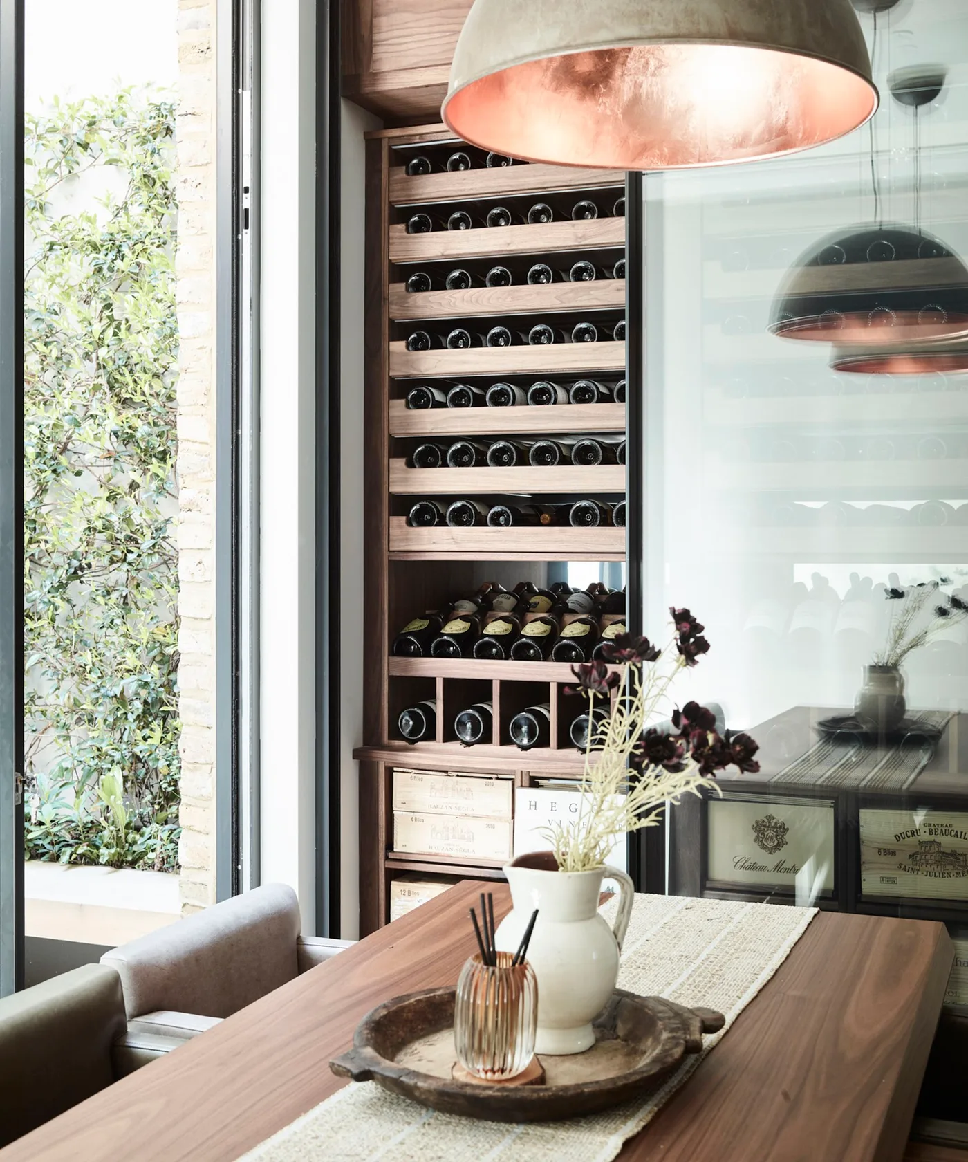 Tall wood bottle-storage compartments beside a wood dining table, pale bench and copper-colored pendant interior