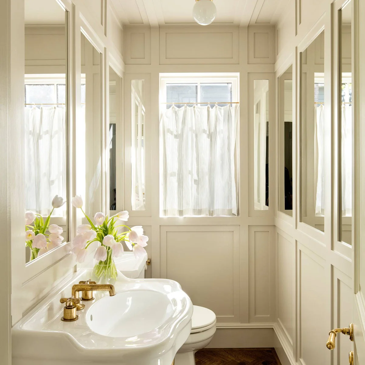 A pale paneled powder room with inset mirrors, a shaped white basin and a short curtain over the lower window.