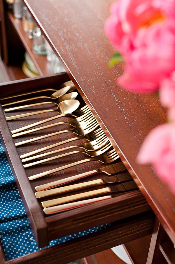 A shallow wood sideboard drawer holds neatly grouped gold-colored cutlery below the cabinet top beside an open storage compartment