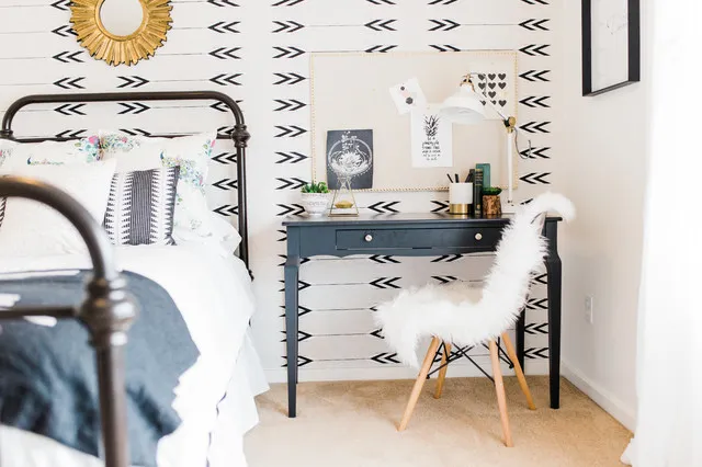 A dark narrow desk and white textured chair stand against a black-and-white arrow-patterned wall beside a dark metal bed and small artwork