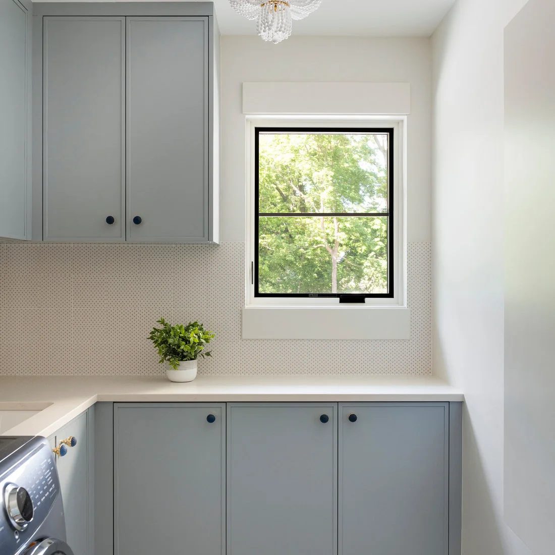 Muted gray cabinets and a white counter sit beneath a small black-framed window and a finely patterned pale backsplash beside a machine.