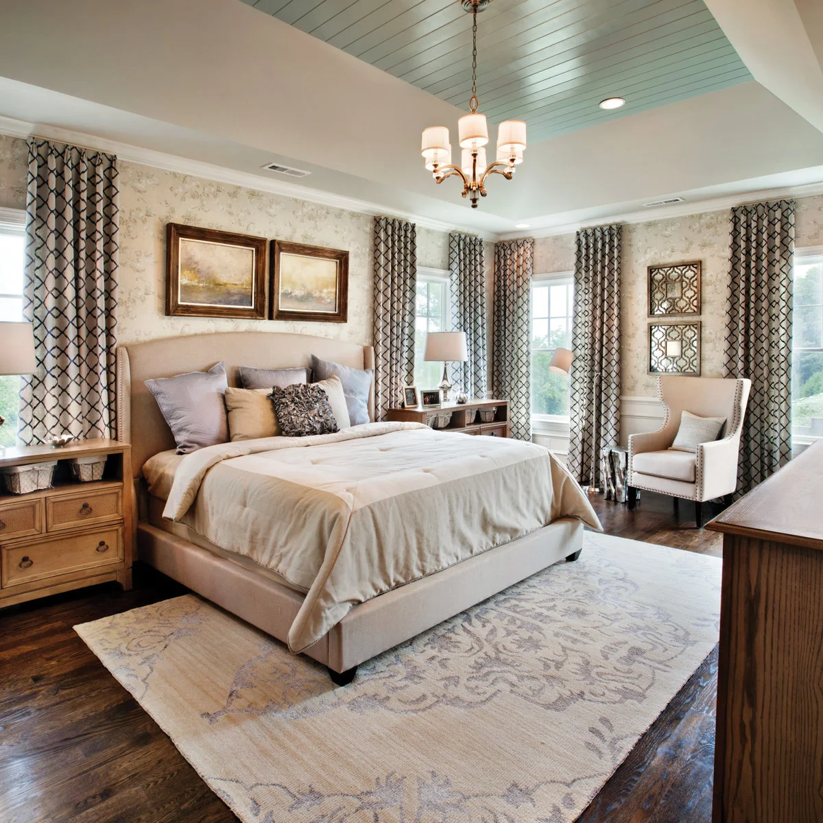 A pale muted patterned rug lies beneath a cream bed surrounded by patterned wallpaper and tall curtains.