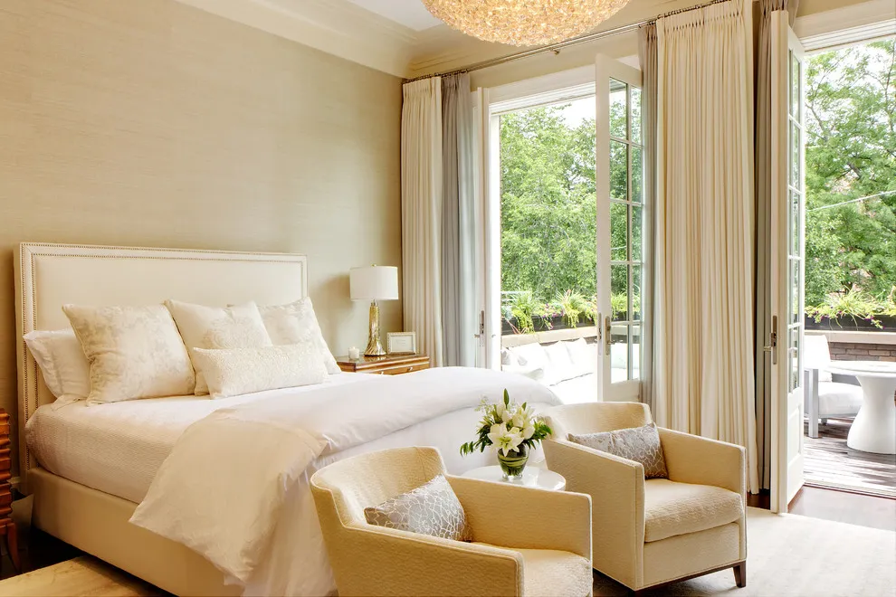 A cream bed sits beside small yellow chairs and tall curtains near broad windows