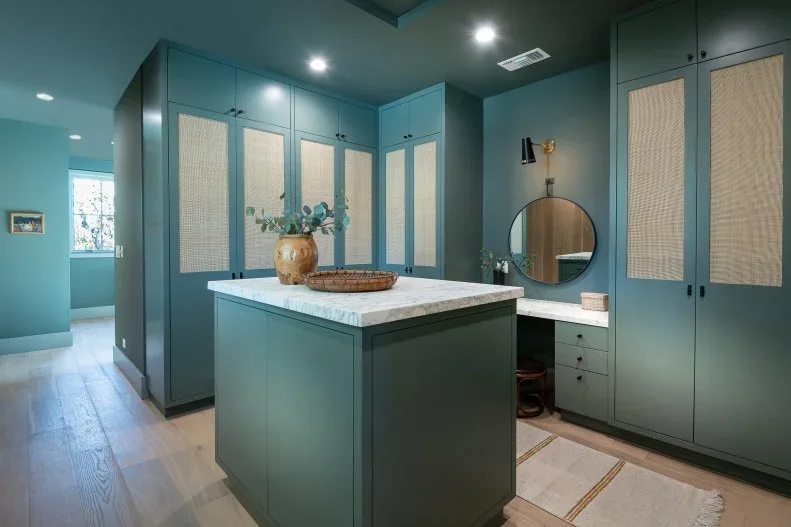 Green closet cabinets with woven door inserts surround an island and a small mirrored dressing space.