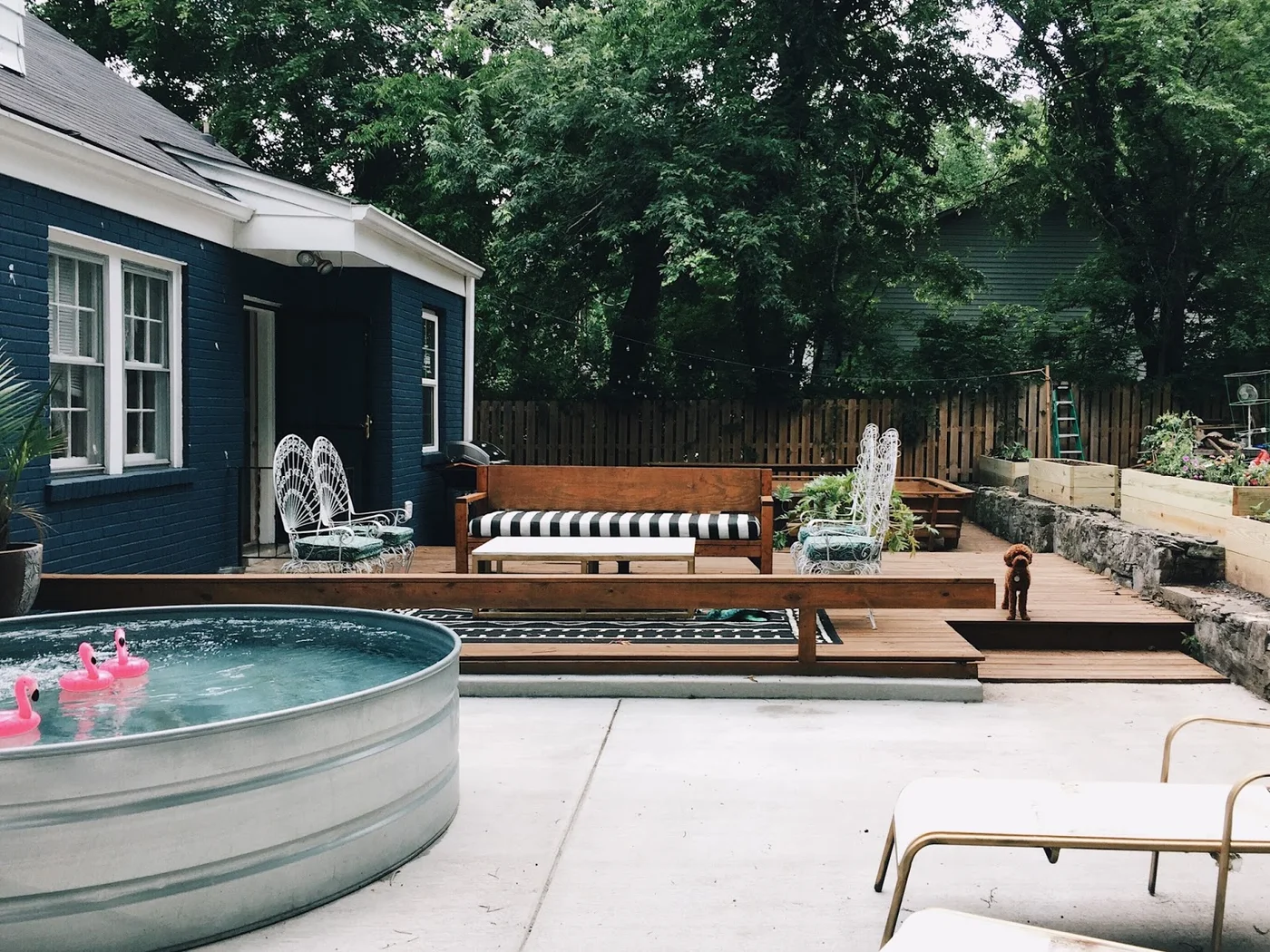 Round metal stock tank pool beside a wood deck with striped cushions, white chairs and planted borders along a stone wall.