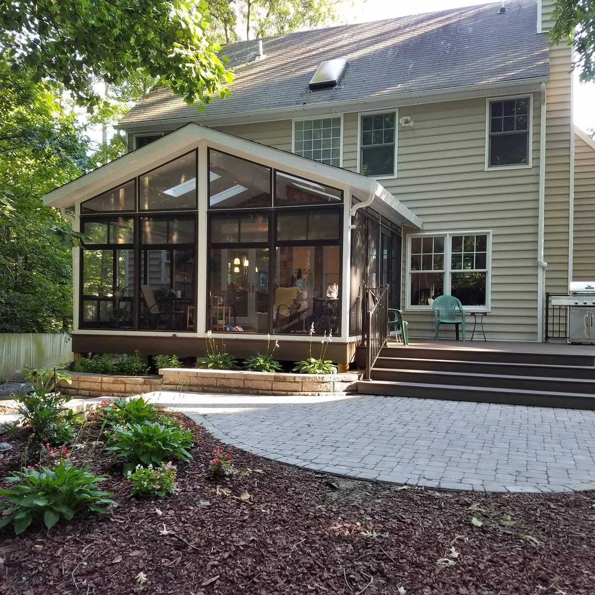 A gabled screened porch attached to a house beside broad deck steps and a paved garden seating area.