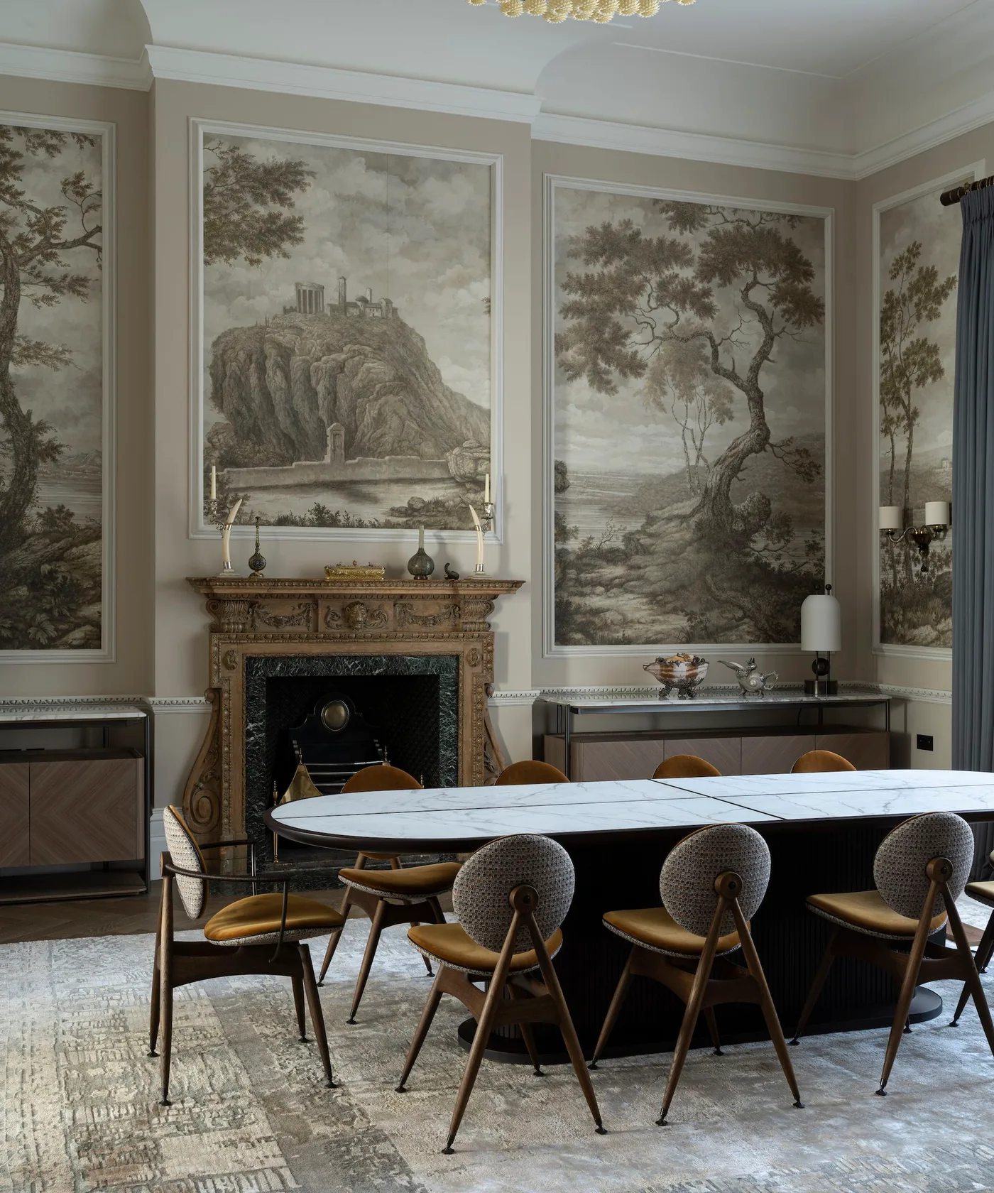 Dining room with framed scenic gray-brown wall panels, a carved fireplace, elongated pale table and rounded gray-backed chairs with ochre seats