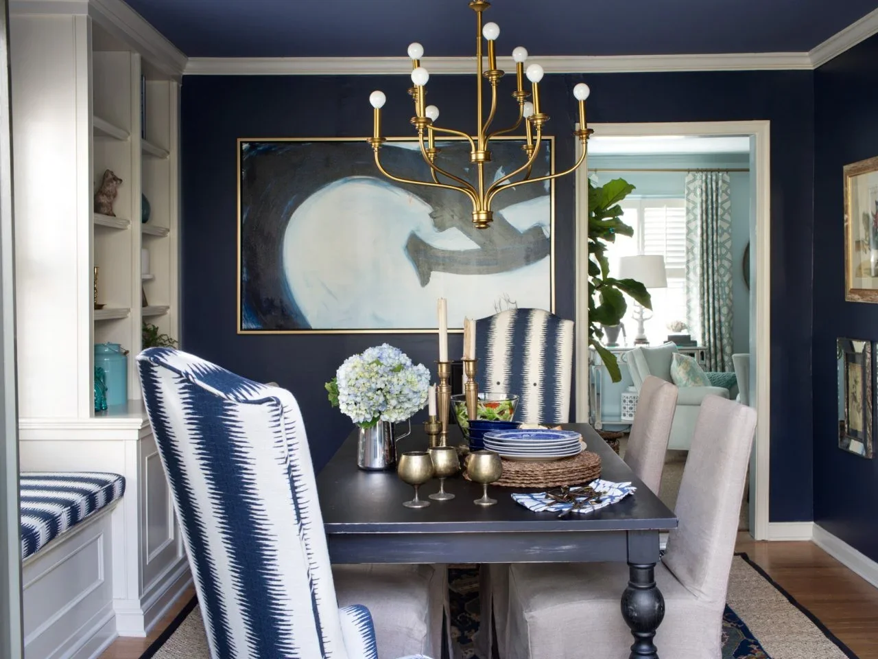 A navy dining room has blue-and-white patterned end chairs, pale side chairs, a dark table and a gold-colored chandelier.