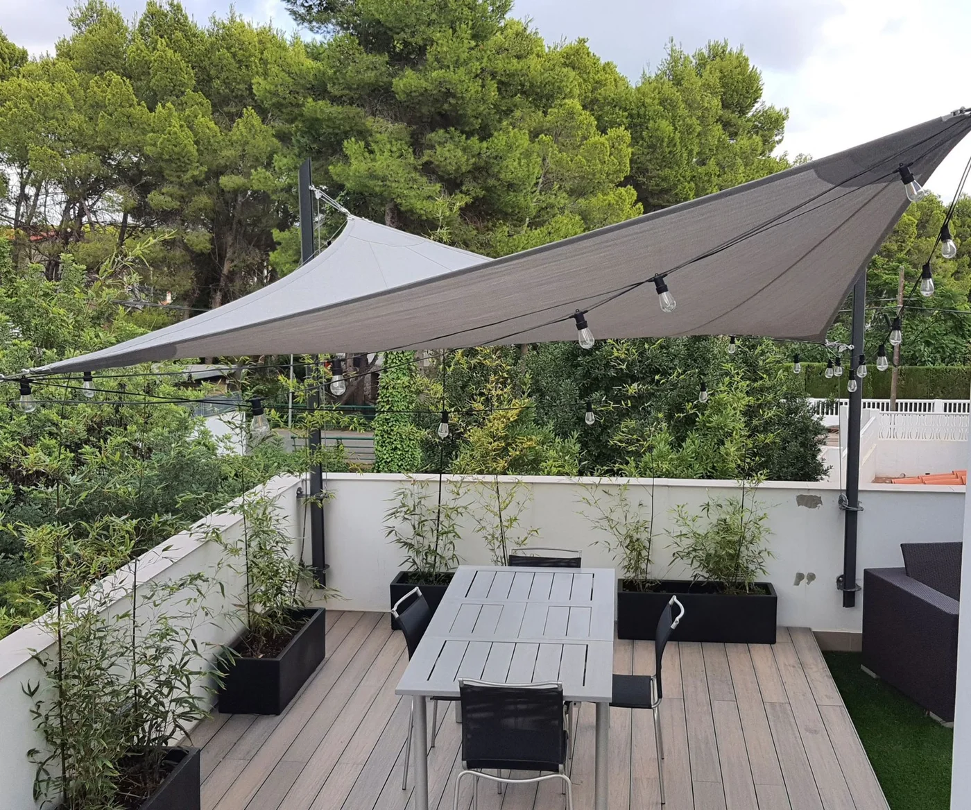 Shade sails and tall container planting surround a rectangular table on a rooftop terrace