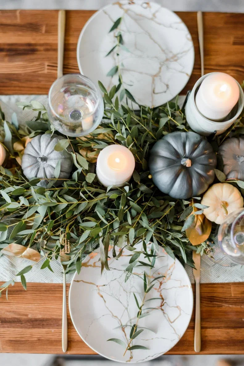 Gray and pale pumpkins sit among long green foliage beside white veined plates and candles on a wooden table