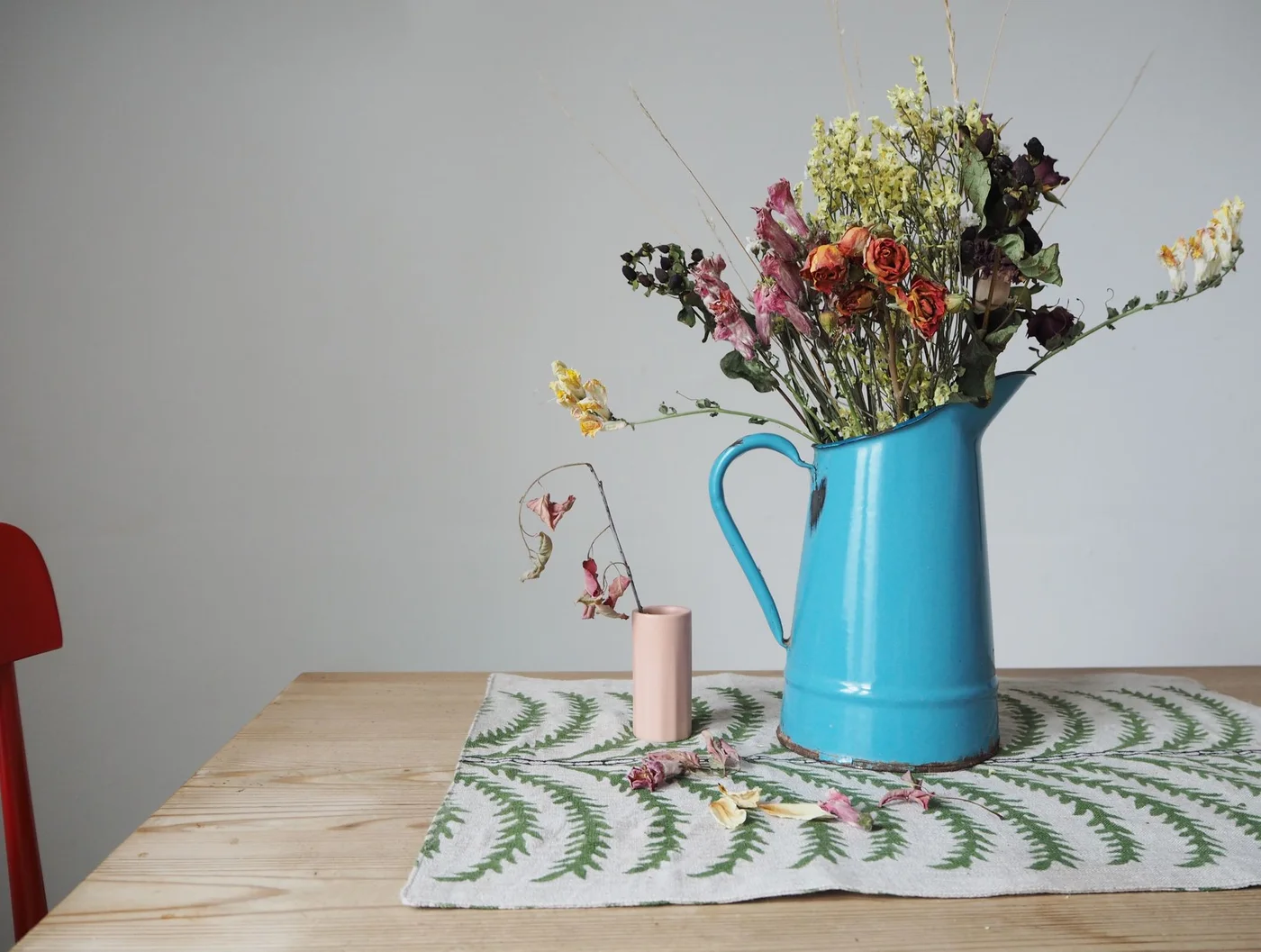 A turquoise jug holds a loose dried bouquet on a pale wood table beside a small pink vase and a fern-patterned cloth.