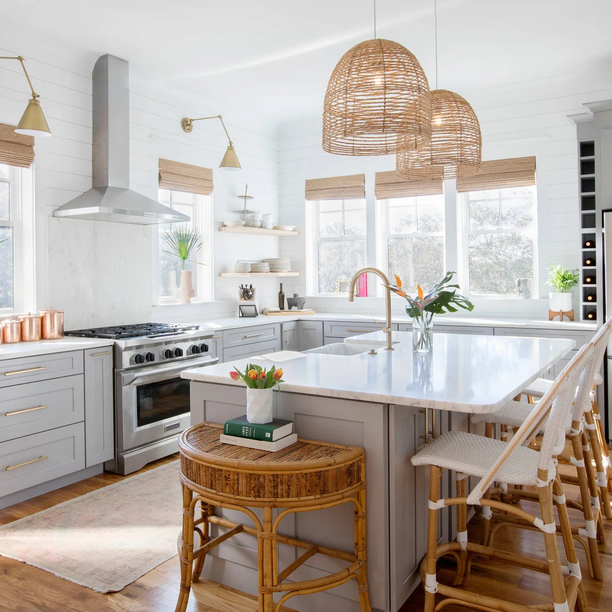 Two broad woven dome pendant shades hang above a pale island beside warm metal wall lights, white wall boards and woven-seat stools.