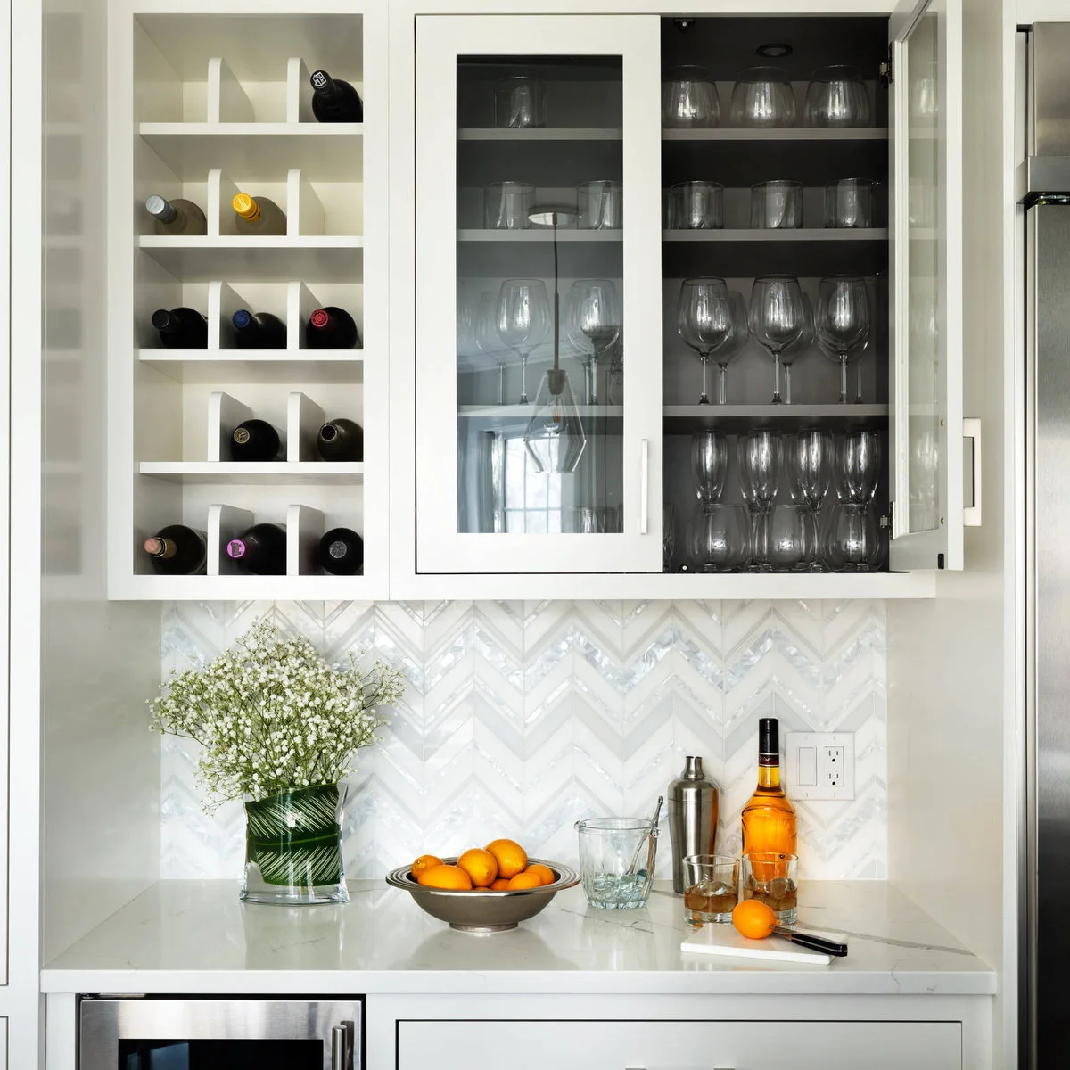 White mini bar with bottle cubbies, dark-backed glass cupboard, herringbone backsplash and clear counter area