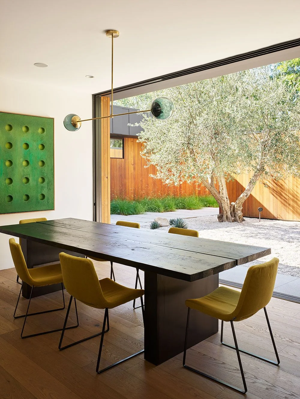 Mustard dining chairs on slim dark metal bases surround a long dark table beside green geometric artwork and broad garden-facing glazing beneath a two-ended pendant