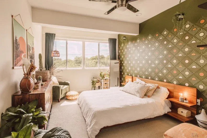 A wood bed with white bedding sits against a green patterned accent wall beside broad windows, a wood dresser, hanging plants and a small chair