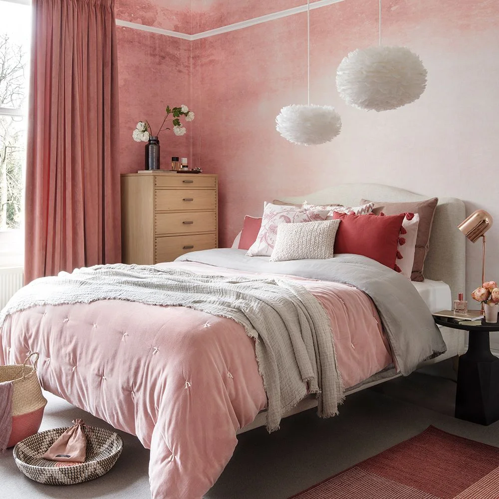 Mottled pink plaster-effect wallpaper surrounds a pale bed with blush bedding, a timber chest and white pendant lights.